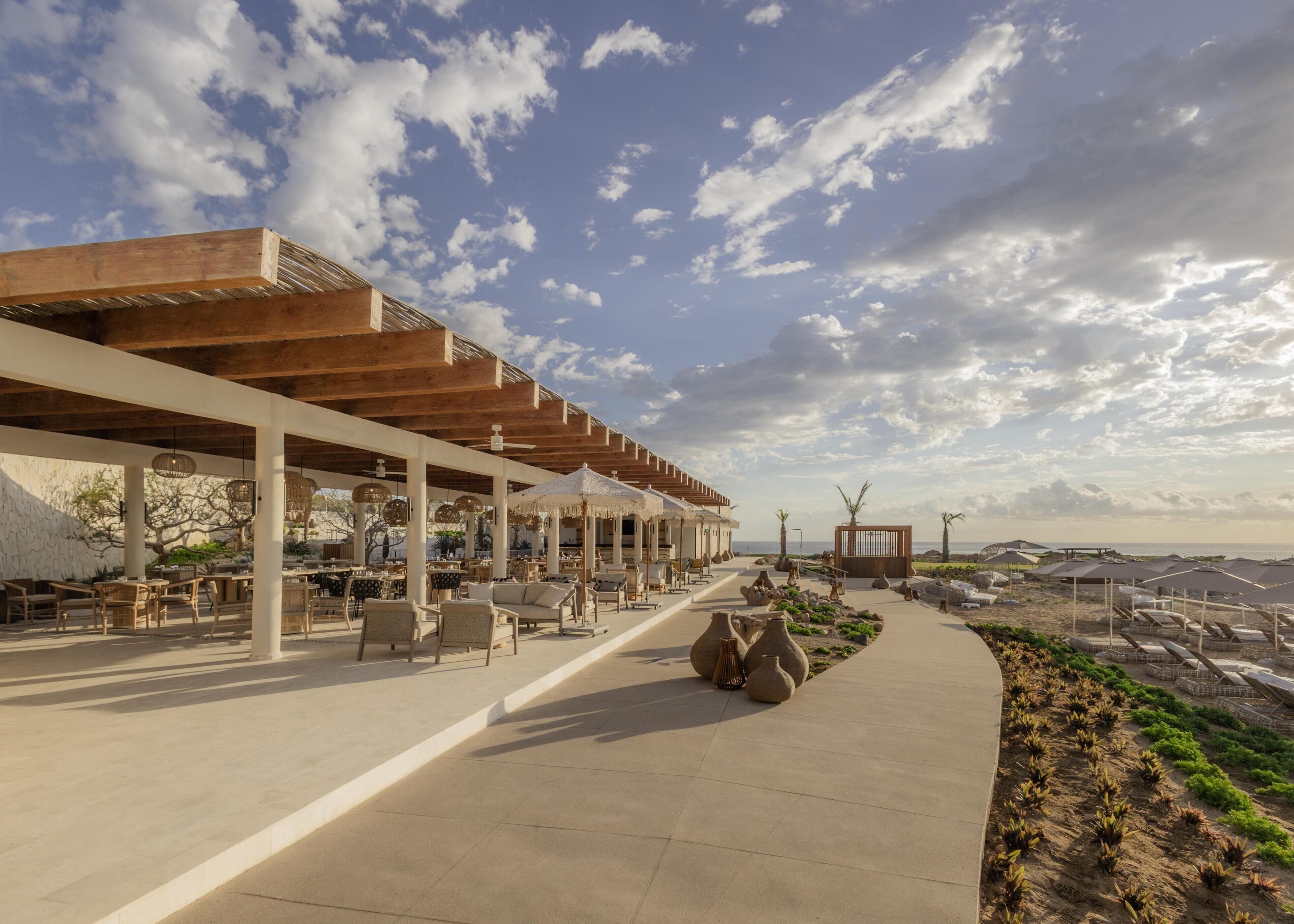 Un espacio al aire libre con mesas y sillas, una sección cubierta con sombrillas y vistas a la playa.