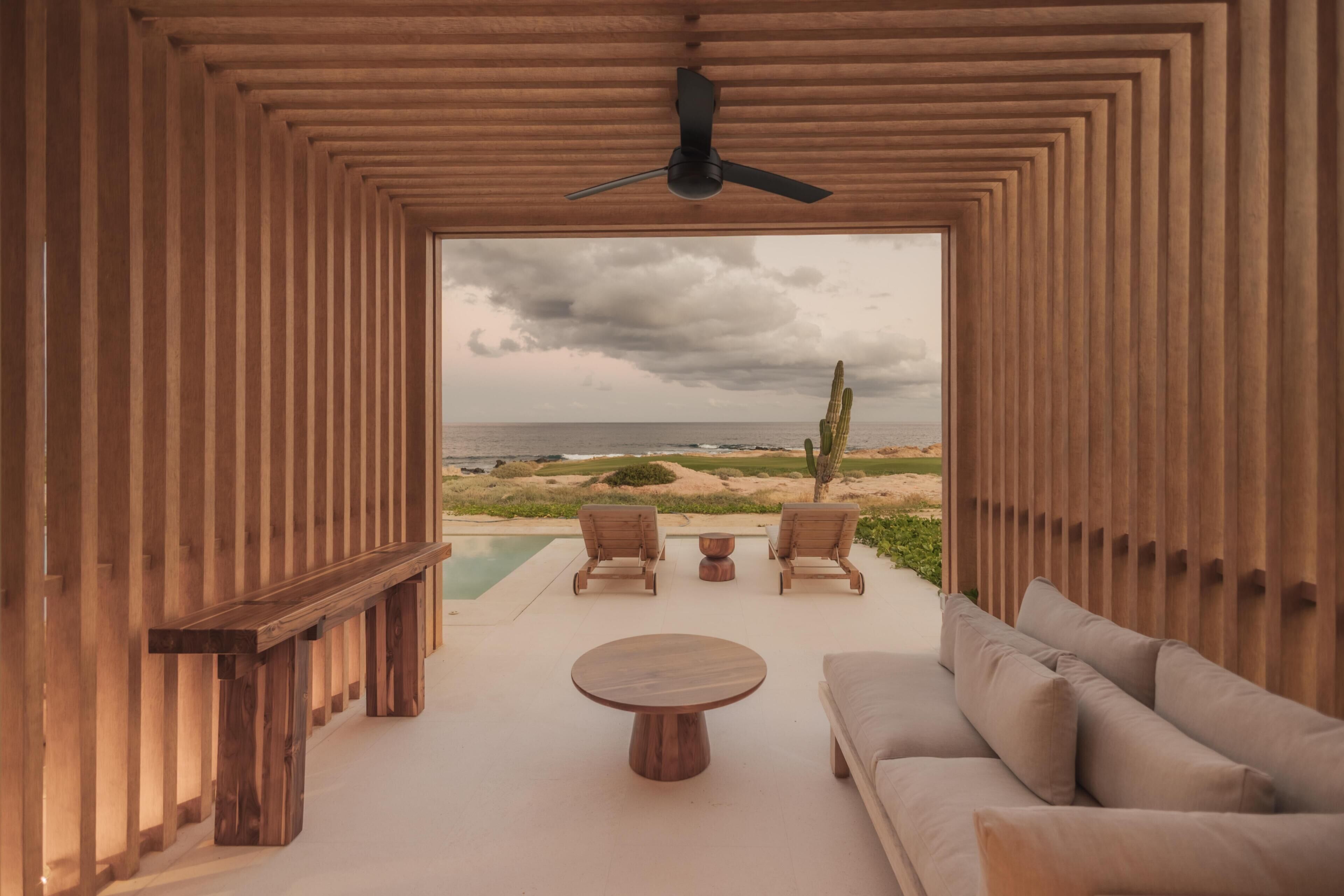 A modern interior with a wooden slatted ceiling, a ceiling fan, a pool, and a view of the beach.