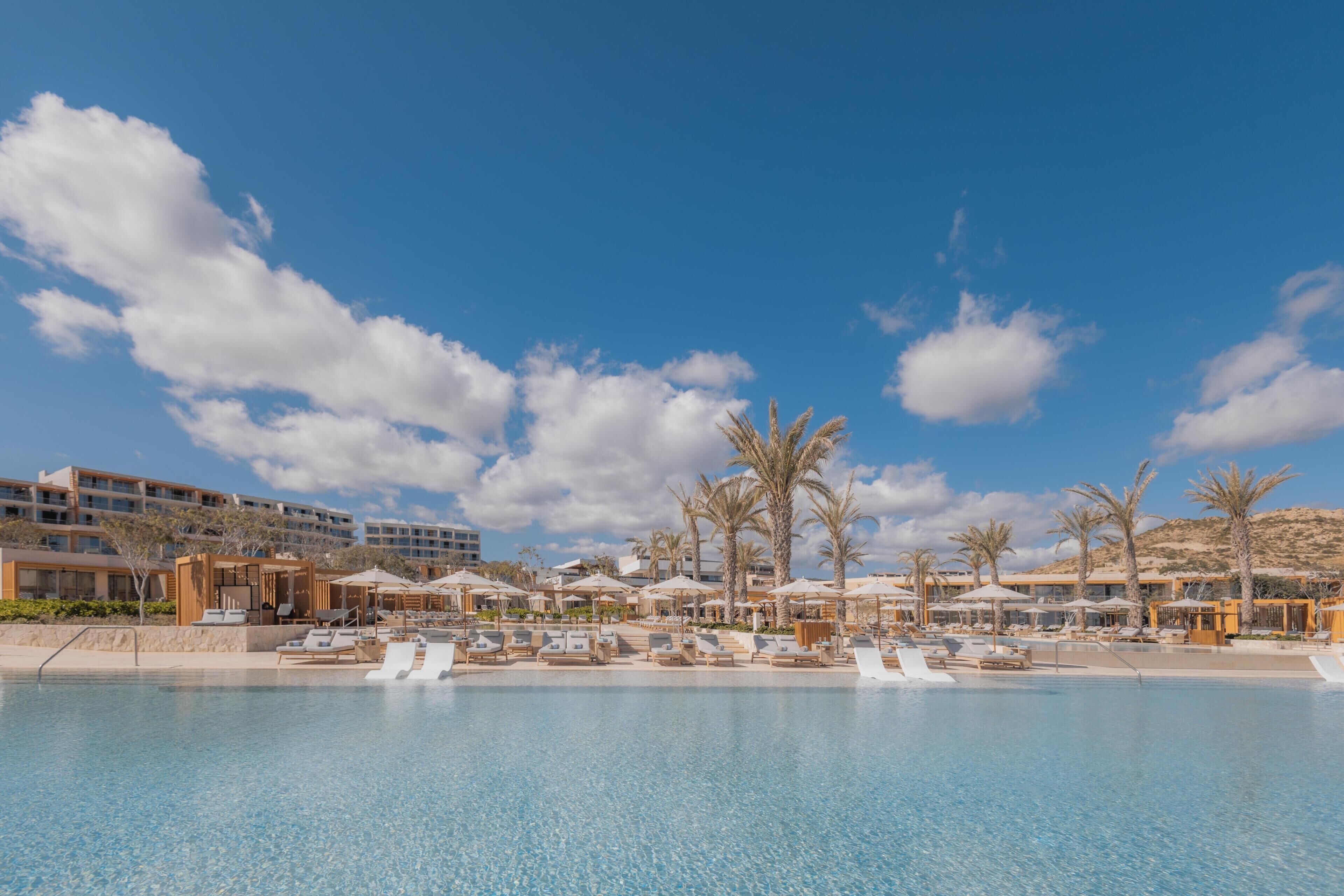 Un área de piscina en un complejo turístico con sillas de descanso y sombrillas, rodeada de palmeras y edificios modernos.