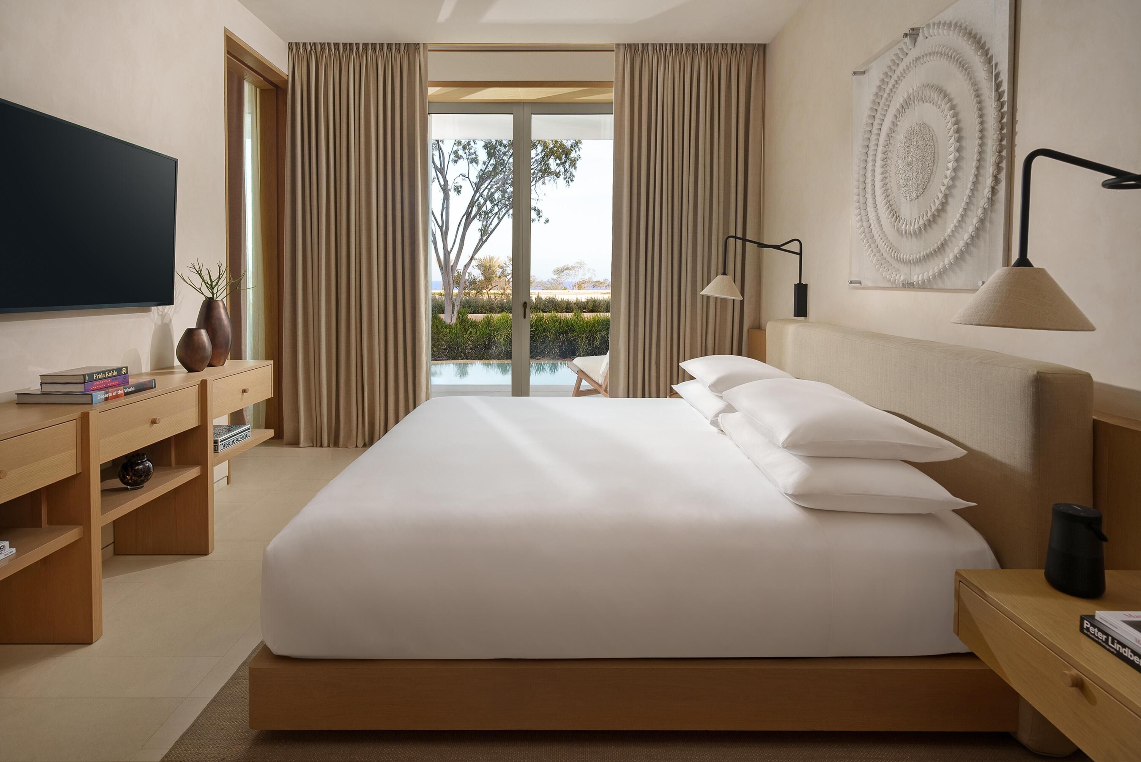 A modern hotel room with a large bed, a flat-screen TV, a desk with books, and a view of a pool and greenery.