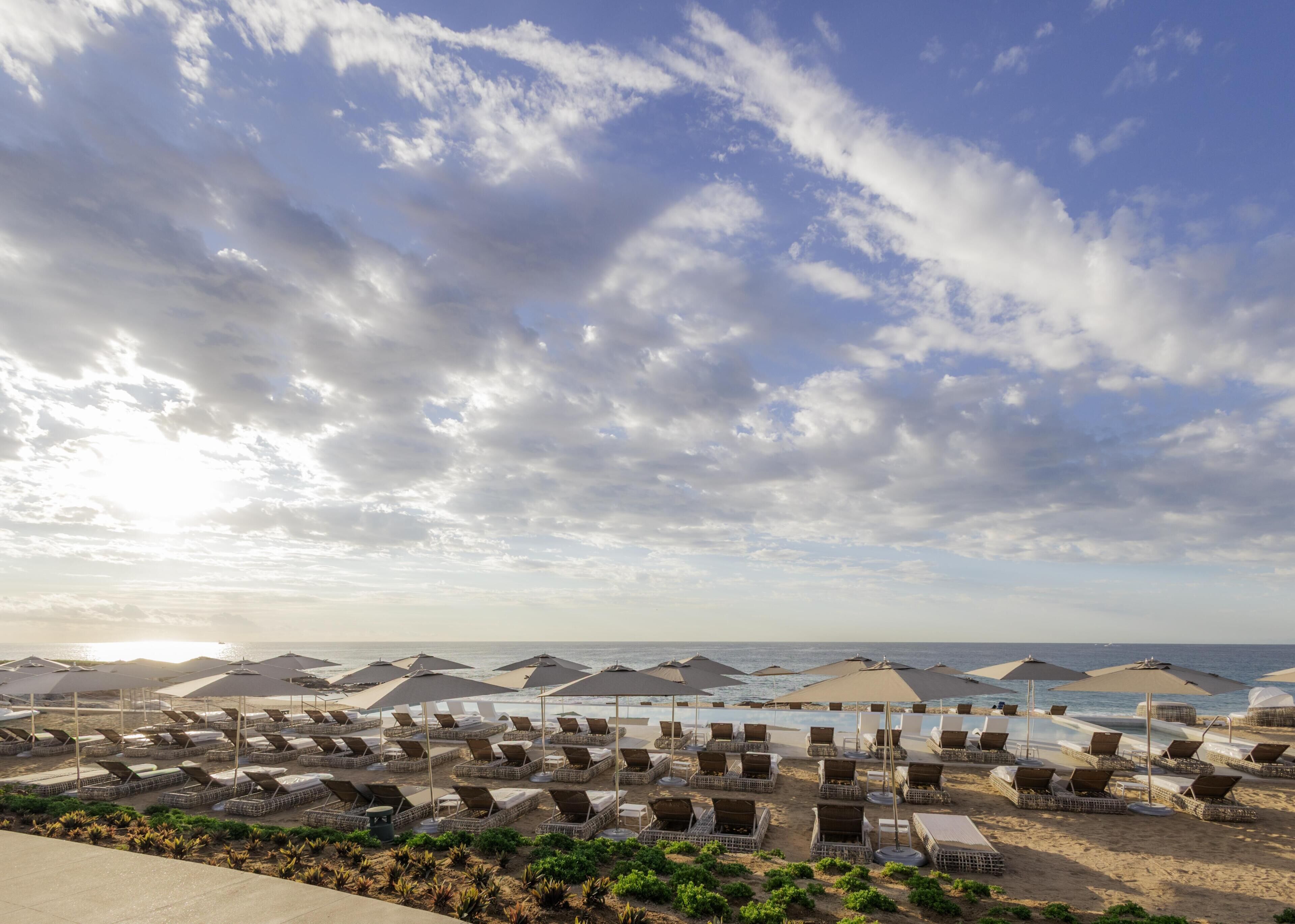 A beachside resort with rows of sun loungers and umbrellas, a clear sky, and the ocean in the background.
