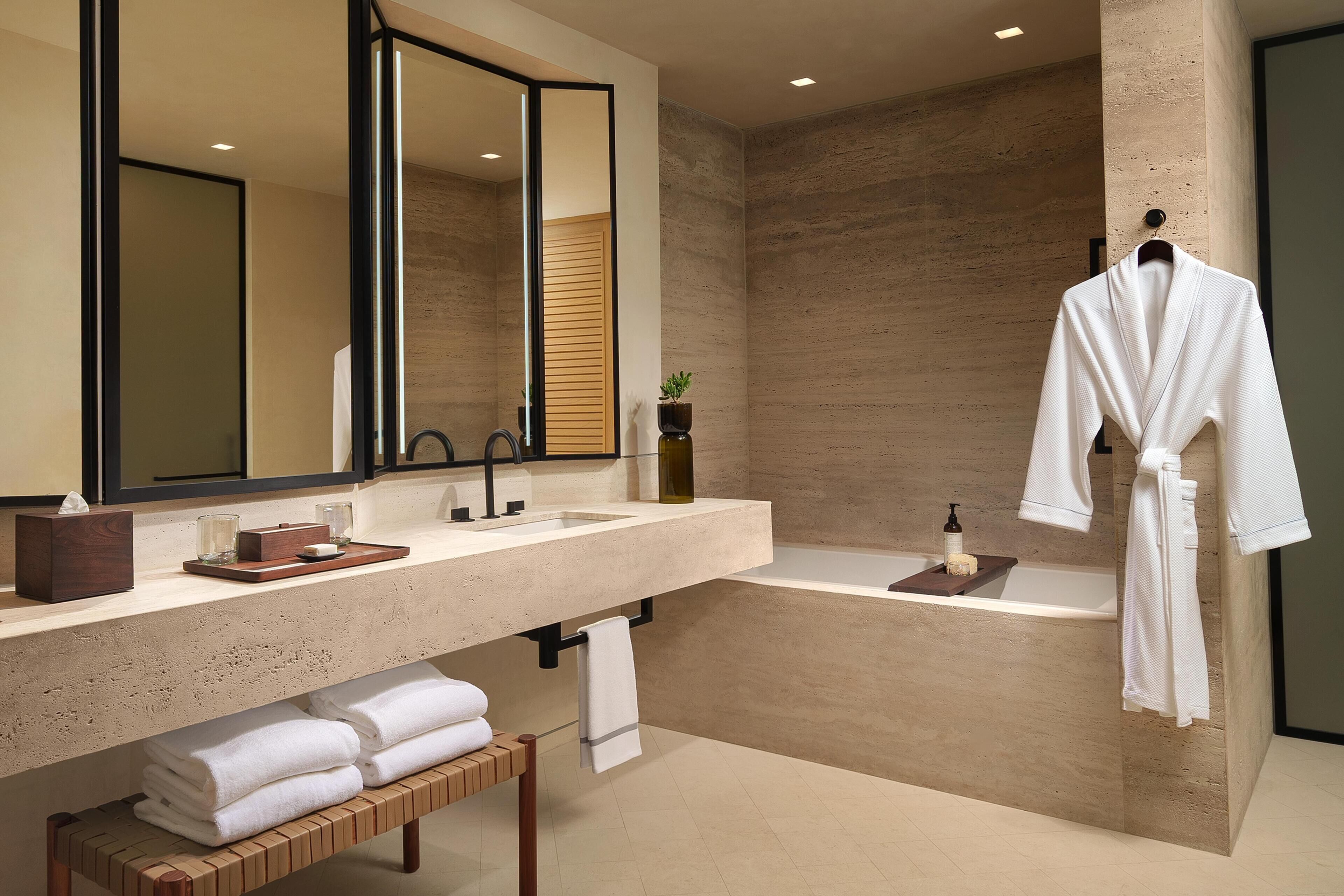 A modern bathroom with a large mirror, a bathtub, and a wooden bench with towels.