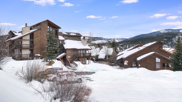 Una cabaña nevada con varias construcciones de madera, una chimenea de piedra y vistas a montañas cubiertas de nieve.