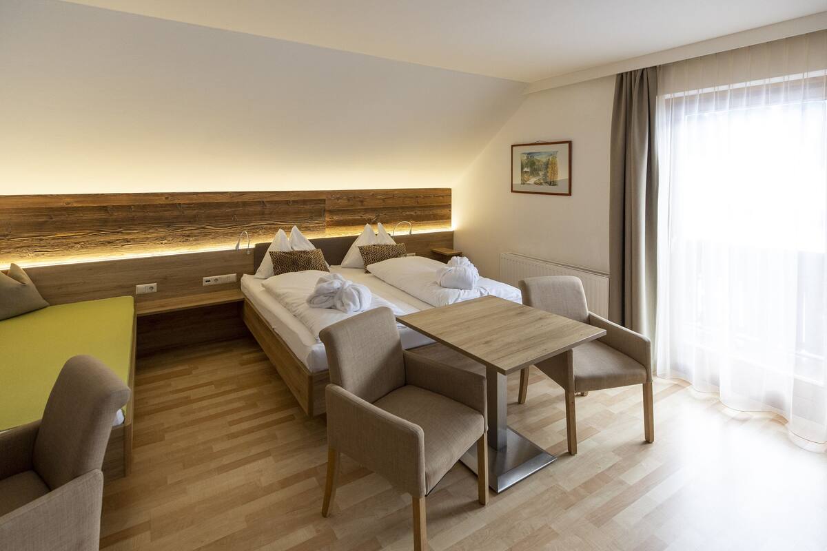 A modern hotel room with a large bed, a wooden headboard, a dining table, and a sofa.