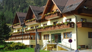 Ein Hotel mit mehreren Balkonen, die mit Blumen geschmückt sind, einer Treppe zum Eingang und einer Aussicht auf eine gebirgige Landschaft im Hintergrund.