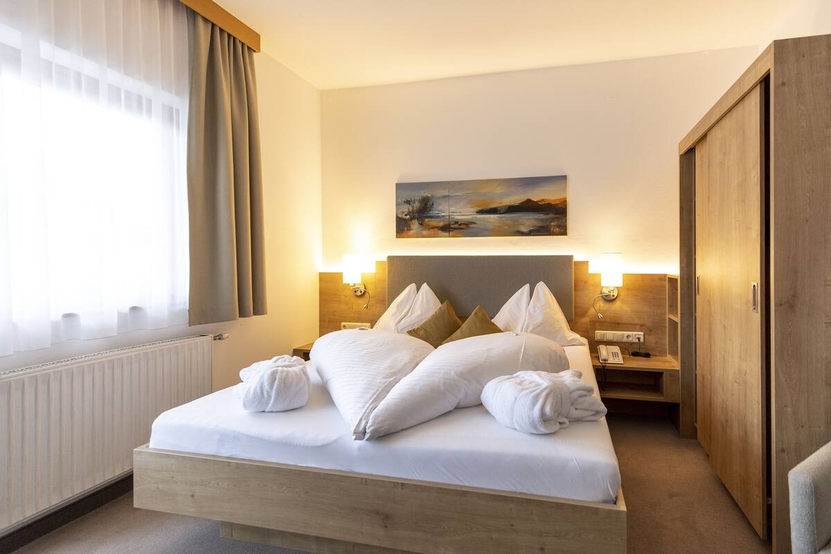A hotel room with a wooden bed, white linens, a painting on the wall, and a nightstand with a lamp.