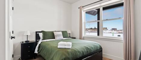 A bedroom with a bed, a nightstand, a window with curtains, and a view of the outside.