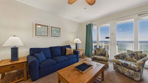 A living room with a sofa, armchair, coffee table, and lamps. There are framed pictures on the wall and a view of the ocean through large windows.