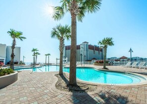 A swimming pool area with palm trees, lounge chairs, and a building in the background. - Ocean Paradise 1425-Ocean Views-Sleeps 7 (Panama City Beach)