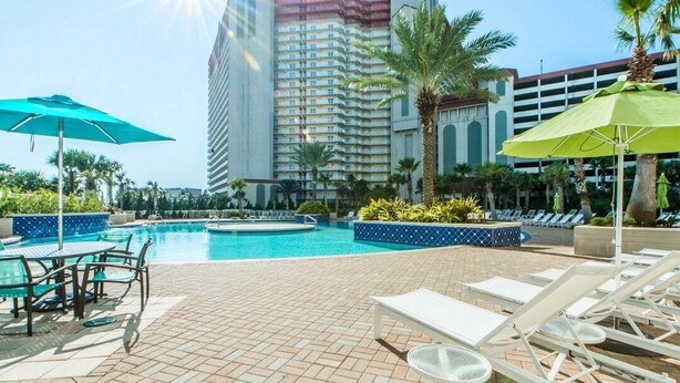 A hotel pool area with lounge chairs, umbrellas, and a large building in the background. - Ocean Paradise 1425-Ocean Views-Sleeps 7 (Panama City Beach)