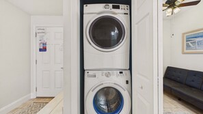 A laundry room with stacked washers and dryers, a door with safety and beach safety signs, a ceiling fan, and a framed picture on the wall. - Ocean Paradise 1425-Ocean Views-Sleeps 7 (Panama City Beach)