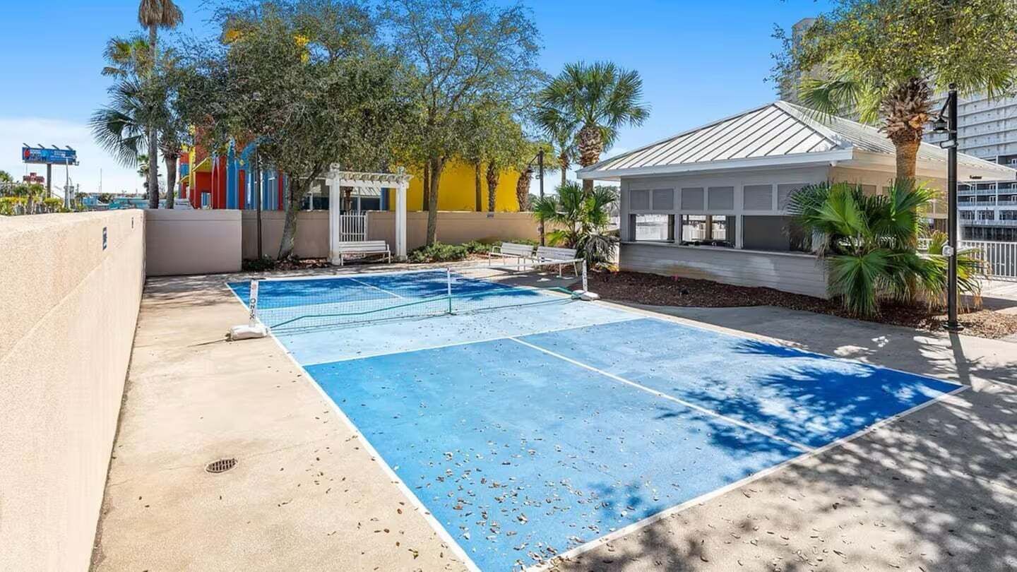 Een tennisbaan met een net, omringd door palmbomen en een gebouw met een wit dak.