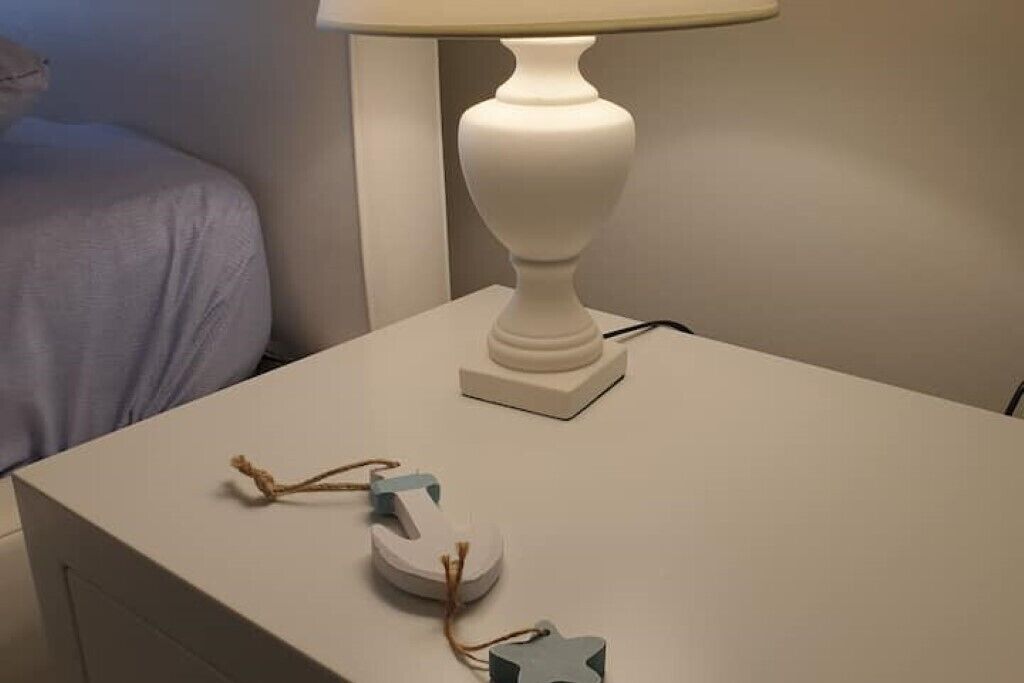 Una mesita de noche blanca con una lámpara blanca y un pequeño adorno encima.
