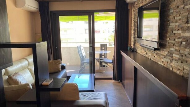 Ein modernes Hotelzimmer mit einer Couch, einem kleinen Tisch, einem an einer Steinwand befestigten Fernseher und einem Balkon mit Blick auf die Stadt.