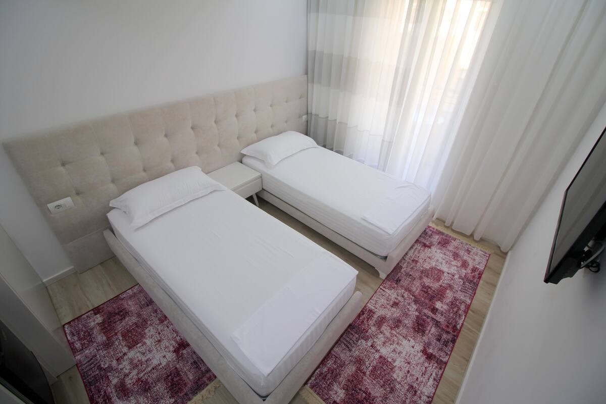 A bedroom with two single beds, a tufted headboard, a red and white patterned rug, and a large mirror on the wall.