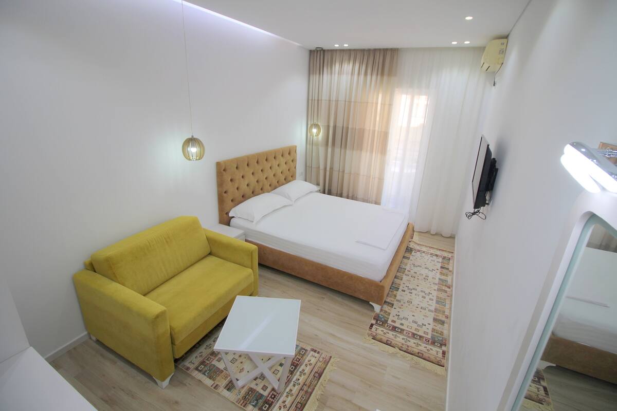 A hotel room with a bed, a yellow sofa, a small white table, and a TV.