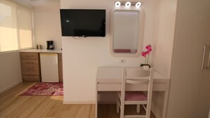 Une chambre avec un téléviseur fixé au mur, un petit bureau avec une machine à café, une coiffeuse blanche avec un miroir et une armoire.