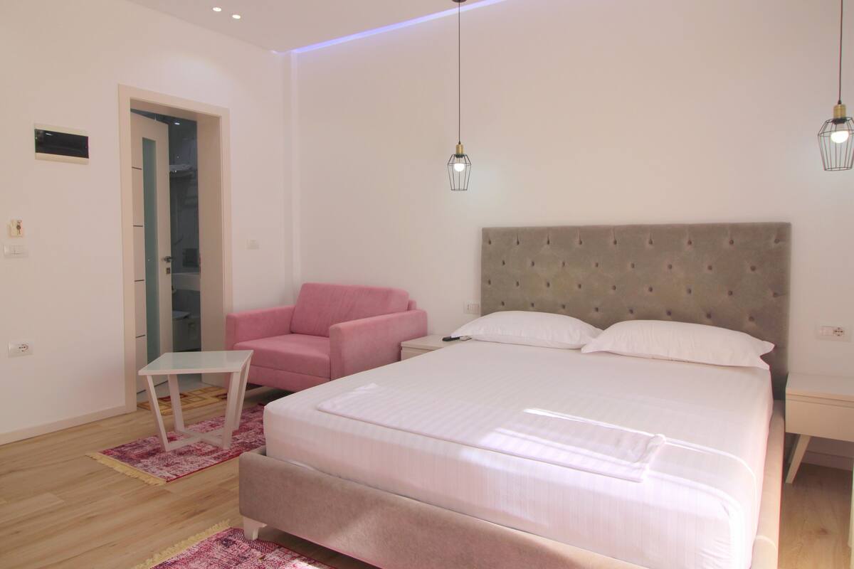 A modern bedroom with a large bed, a pink chair, a small table, and a door leading to another room.