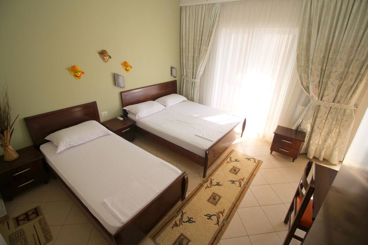 A hotel room with two single beds, a wooden nightstand, a bedside table, a chair, a small table, and a decorative wall piece.