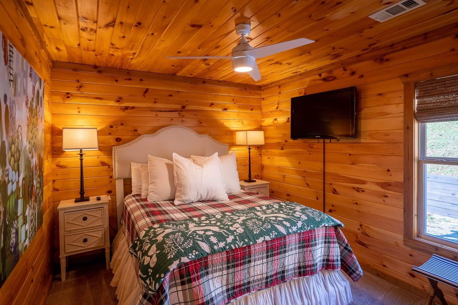 Un dormitorio acogedor con techo de madera, una cama con colcha a cuadros, dos lámparas de noche, un ventilador de techo y un televisor montado en la pared.