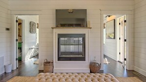 A spacious living room with a fireplace, a large cushioned bench, and a view of the adjacent hallway. - Magnolia Cottage at Julep Farms (Dillard)