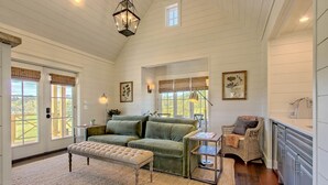 A spacious living room with a green sofa, a bench, and a wicker chair. There are large windows with curtains, a chandelier, and artwork on the walls. - Magnolia Cottage at Julep Farms (Dillard)