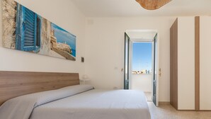 Une chambre à coucher avec un lit, un tableau, une tête de lit en bois, une porte donnant sur un balcon et une vue sur la ville.