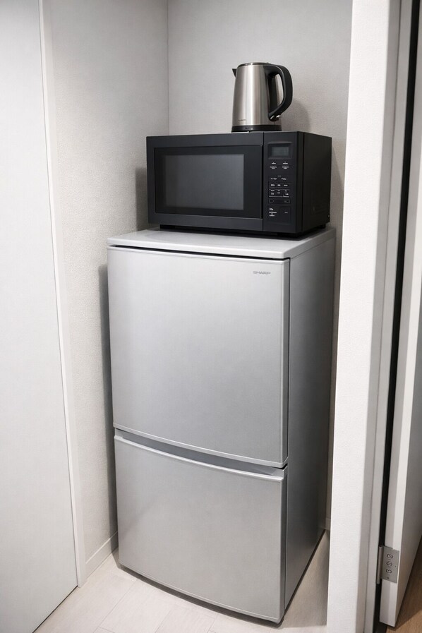 A compact, silver refrigerator with a black microwave on top, situated in a narrow space with a white wall and a wooden floor. - RAHAINA YUHIGAOKA (Osaka)