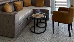 Un salon avec un canapé d’angle gris, une table basse ronde et un fauteuil marron. Trois éléments décoratifs ornent les murs, et une fenêtre est équipée de stores.