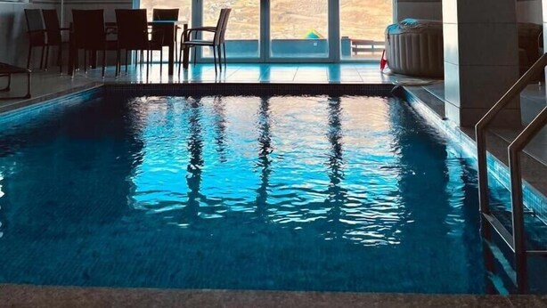 Une piscine intérieure offrant une vue dégagée sur l’extérieur, entourée d’espaces pour s’asseoir et dotée d’un sol carrelé.
