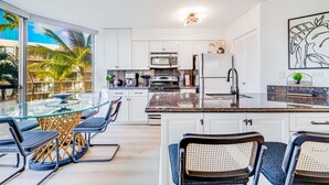 Una cocina moderna con armarios blancos, encimera de granito y comedor con mesa y sillas de vidrio.