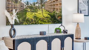 Una habitación con un gran ventanal que da a un patio con palmeras y una piscina. Hay una consola decorativa con objetos decorativos y un cuadro en la pared.