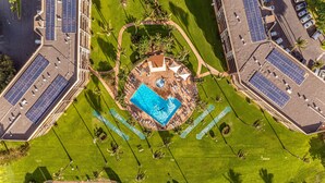 Vista aérea de un resort con piscina central, rodeado de paneles solares, palmeras y un estacionamiento.