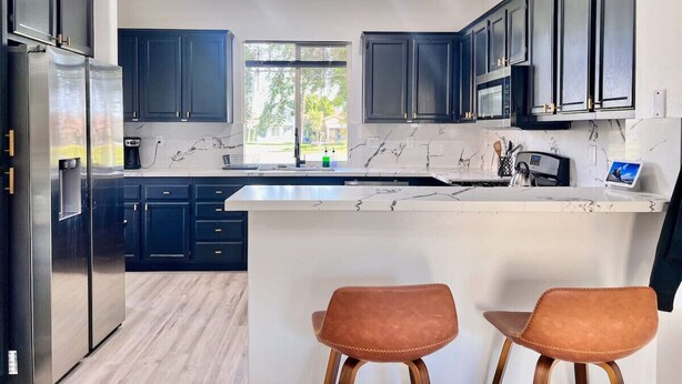 Una cocina moderna con armarios oscuros, un refrigerador de acero inoxidable, una encimera de mármol y dos taburetes.