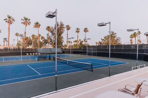 An outdoor tennis court with a net, surrounded by palm trees and street lamps.