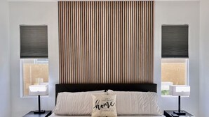 Un dormitorio con una cama grande, dos mesitas de noche con lámparas, un cabecero con listones verticales y ventanas con persianas.