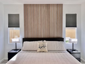 A bedroom with a large bed, two bedside tables with lamps, a headboard with vertical slats, and windows with blinds.