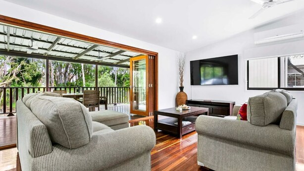 A spacious living area with a wooden floor, two grey armchairs, a coffee table, and a TV on the wall.