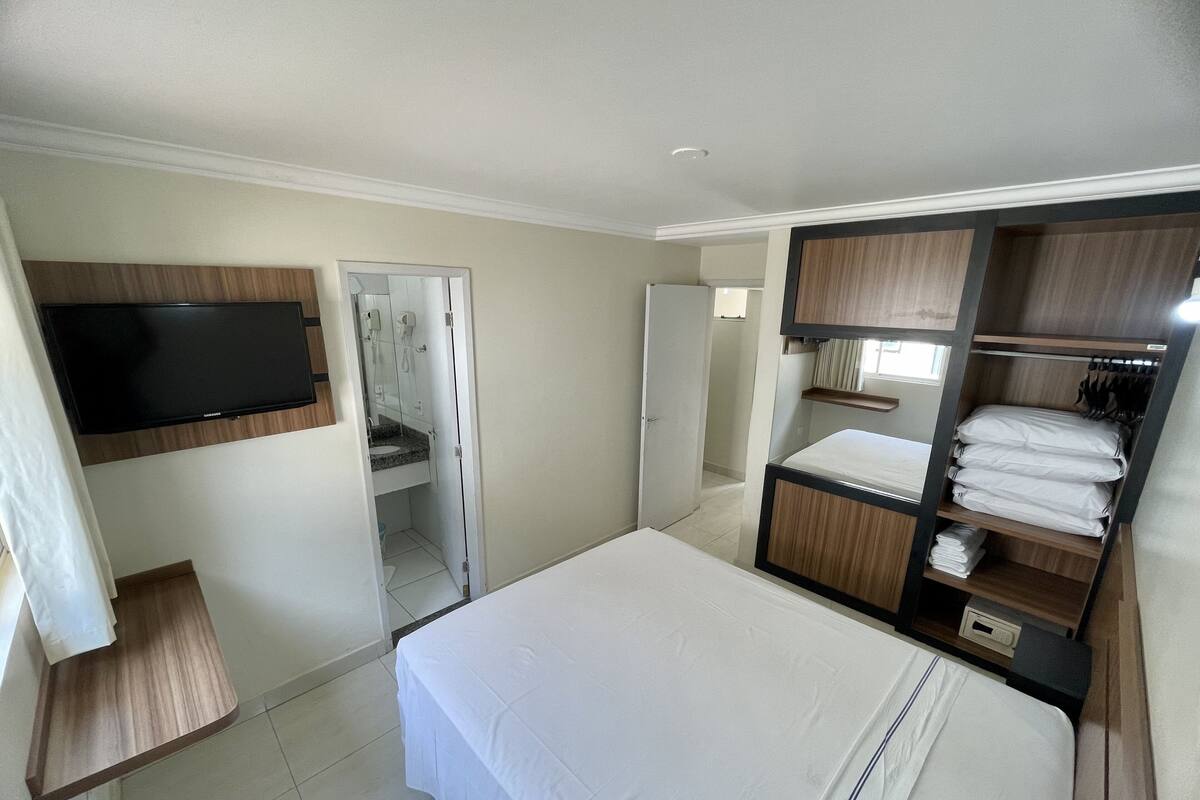 A hotel room with a bed, a TV mounted on the wall, a wardrobe, and a bathroom visible through an open door.