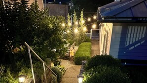 A pathway leading to a house with a sloped roof, surrounded by greenery and illuminated by string lights.
