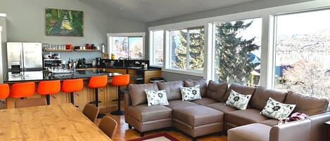 A modern kitchen with bar stools, a dining table, and a large window with a view of trees.