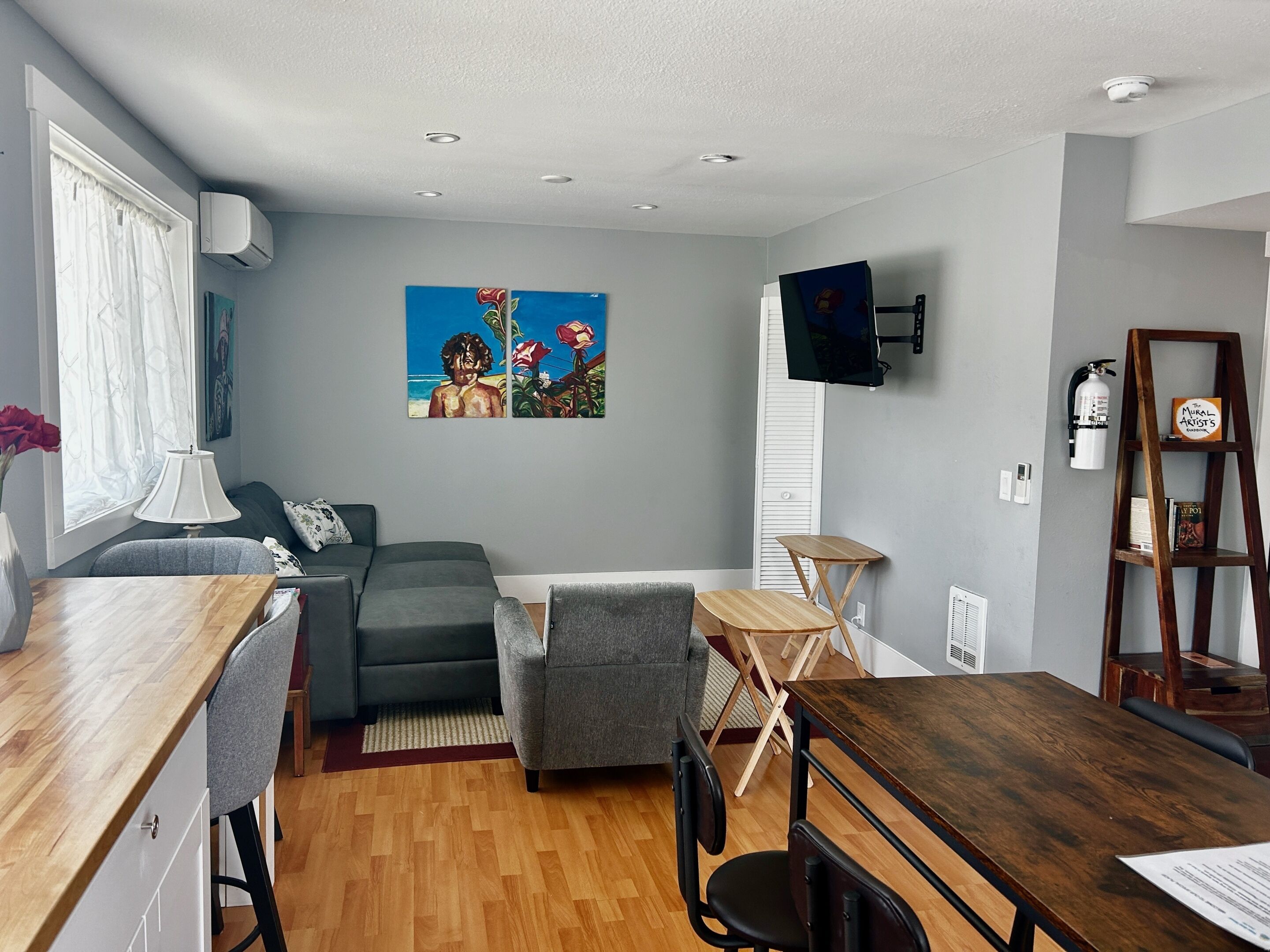 A modern living room with a grey sofa, a wooden dining table, a TV mounted on the wall, and a painting above the sofa.