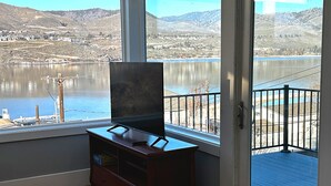 A room with a large window overlooking a lake and hills, a wooden dresser, and a television on a stand. - Water Street Retreat (Chelan)