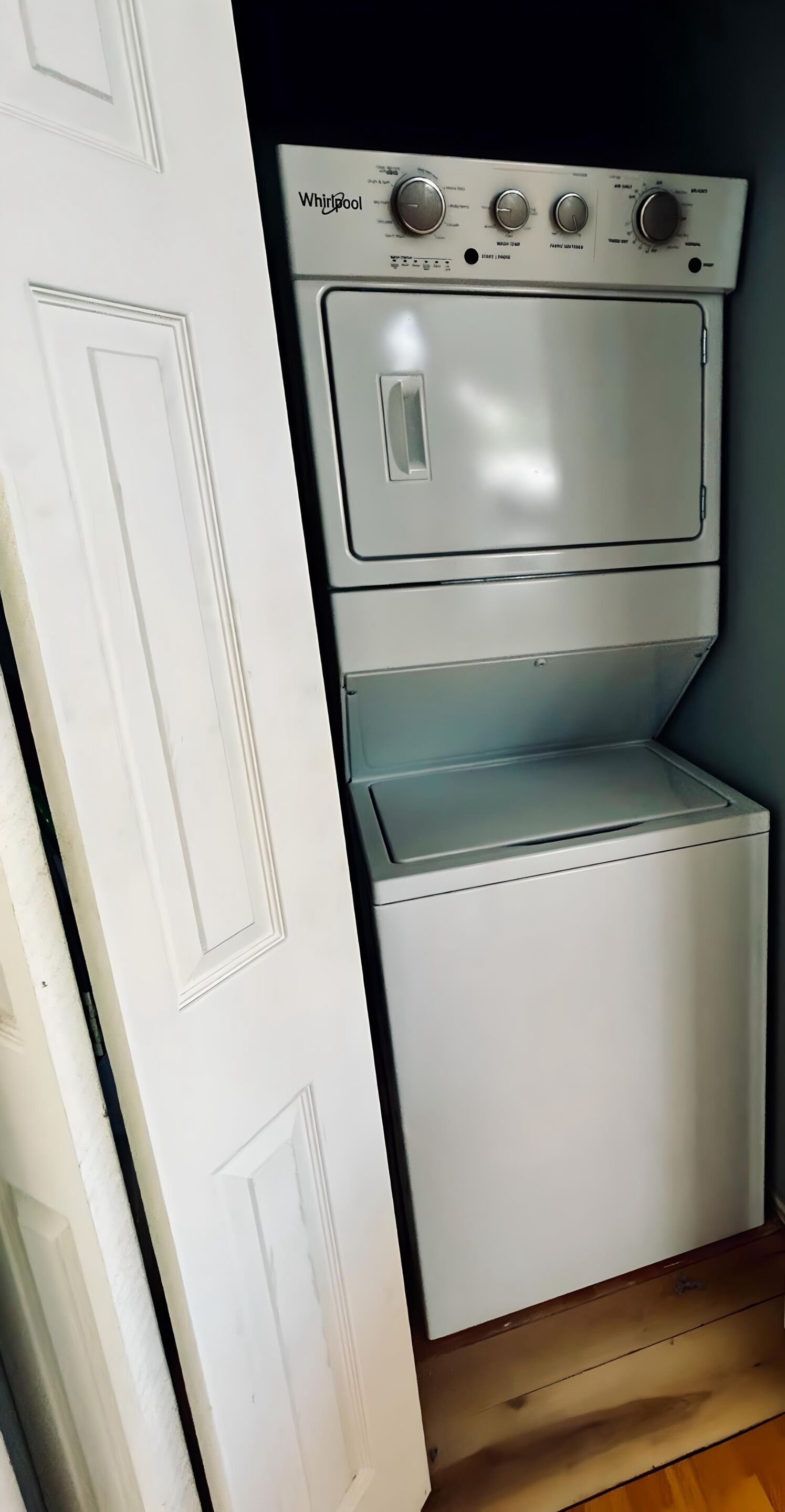 A compact, white Whirlpool stacked washer and dryer unit in a room with wooden flooring and a white door.