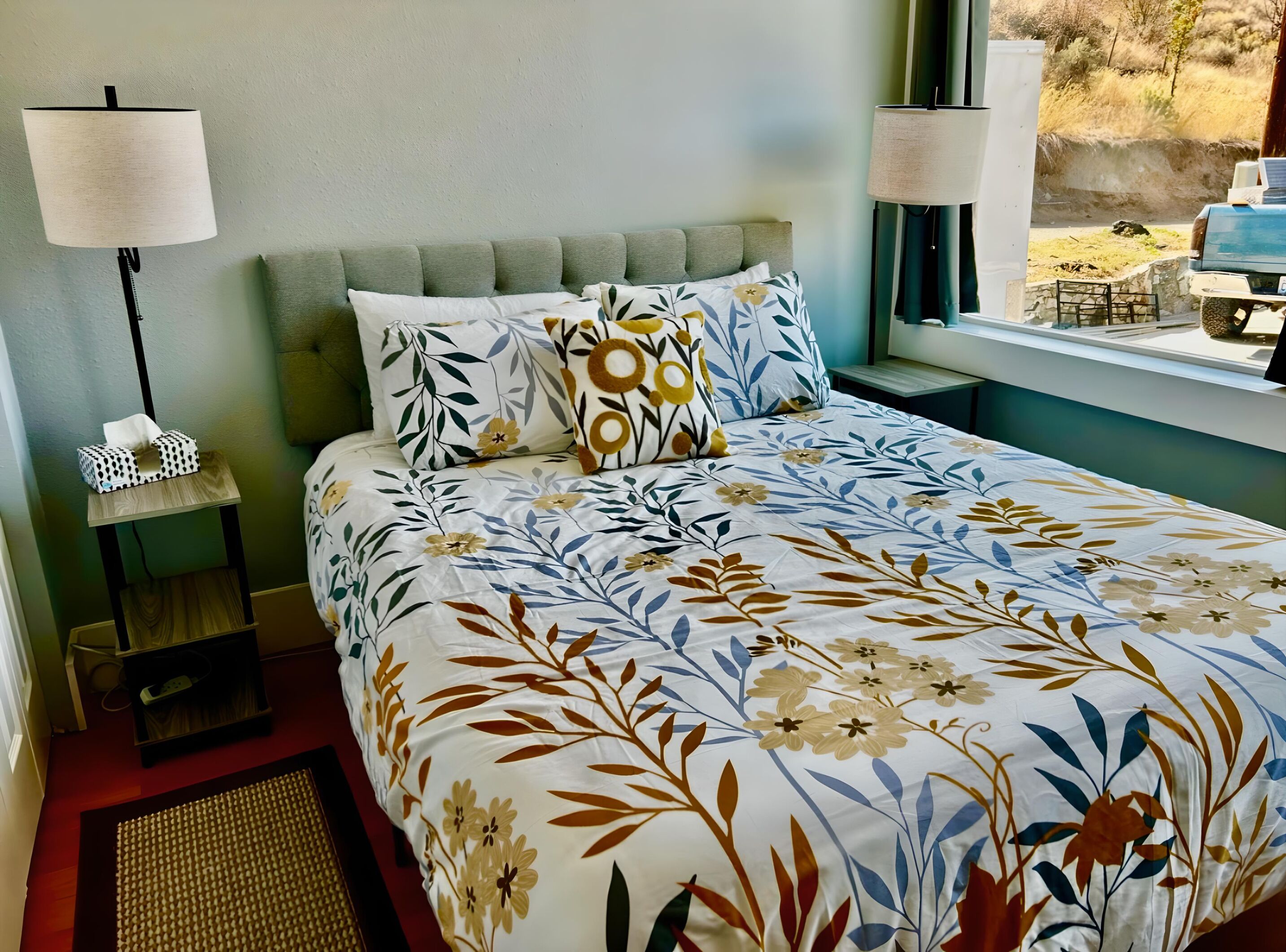 A bedroom with a floral bedspread, a grey headboard, two bedside lamps, and a window with a view of the outdoors.