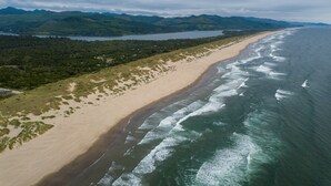 Eine Vogelperspektive zeigt einen Strand mit einer weiten Sandfläche, Wellen und einen bewaldeten Bereich mit Bergen im Hintergrund.