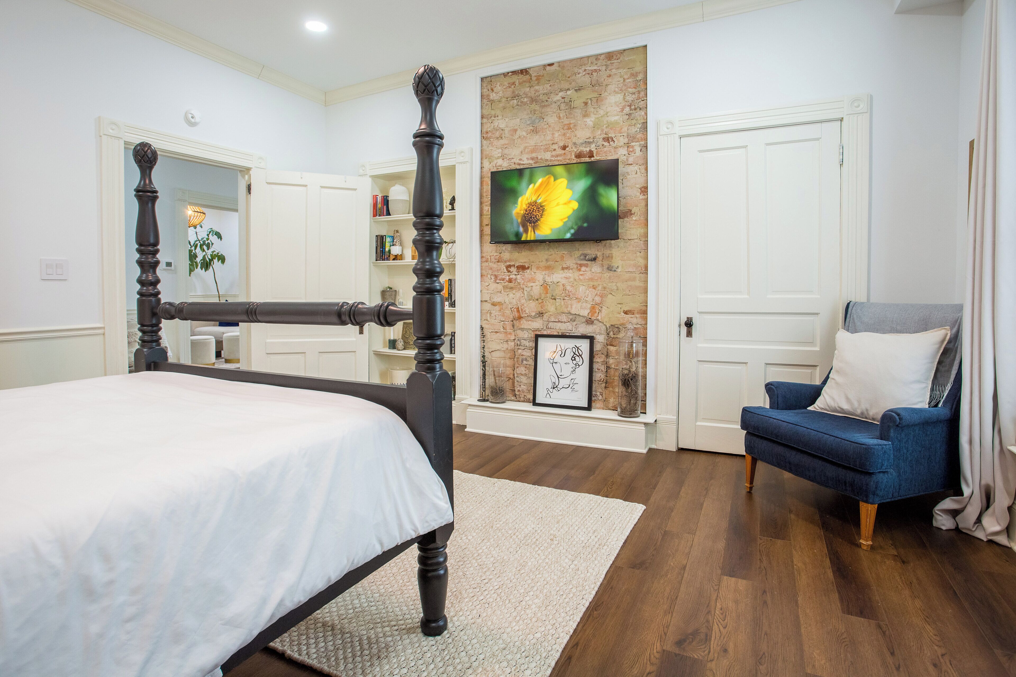 A bedroom with a four-poster bed, a blue armchair, a fireplace, and a TV mounted on a brick wall.
