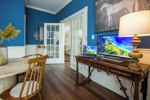 Et værelse med et træskrivebord, en bærbar computer, en stationær skærm og en lampe. Der er en stol og et spisebord med en vase med tørrede planter.