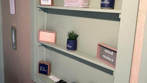 Eine Wand mit Regalen, auf denen verschiedene Gegenstände stehen, darunter Topfpflanzen, gerahmte Zitate und ein kleiner Schreibtisch mit einer Tastatur und einem blauen Objekt.