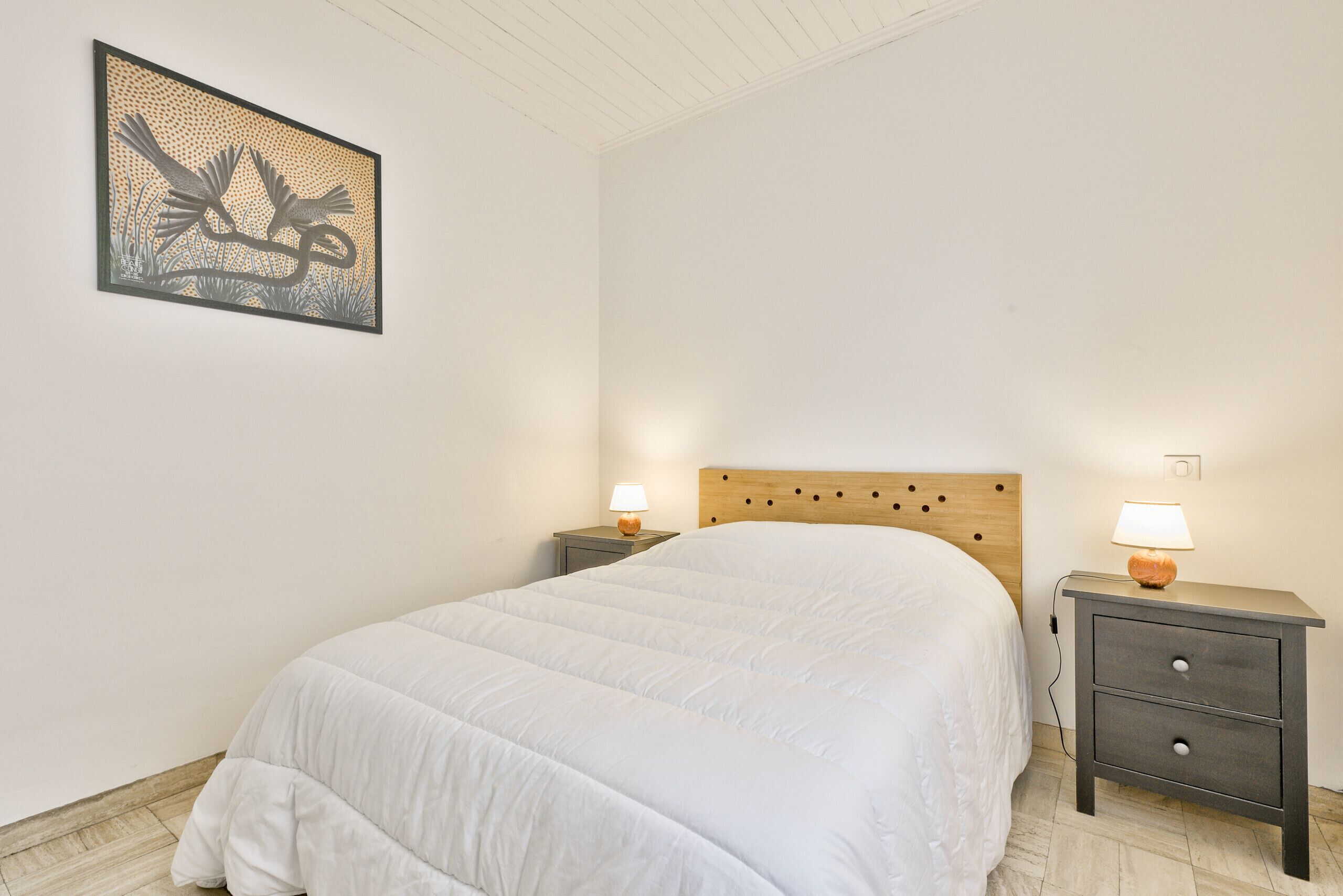 Een slaapkamer met een groot bed, twee nachtkastjes met lampen, een houten hoofdbord en een ingelijst kunstwerk aan de muur.